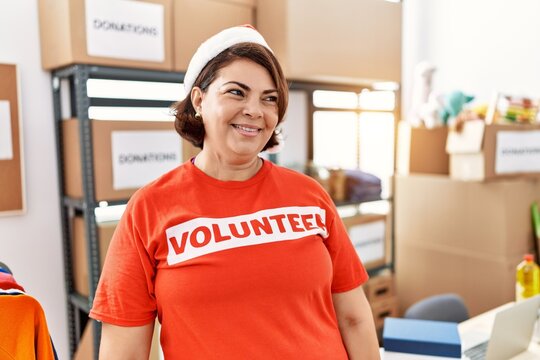 Middle age hispanic woman wearing volunteer t shirt and christmas hat looking away to side with smile on face, natural expression. laughing confident.
