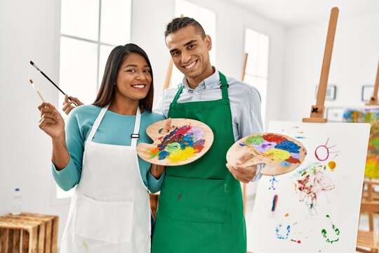 Young latin painter couple smiling happy holding paintbrush and palette at art studio