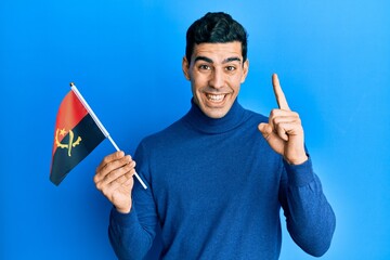 Handsome hispanic man holding angola flag smiling with an idea or question pointing finger with happy face, number one