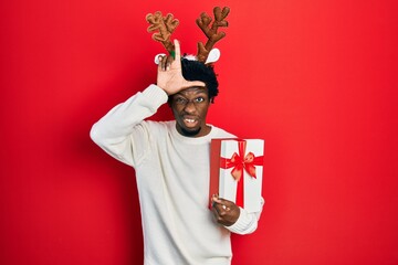 Young african american man wearing deer christmas hat holding gift making fun of people with...