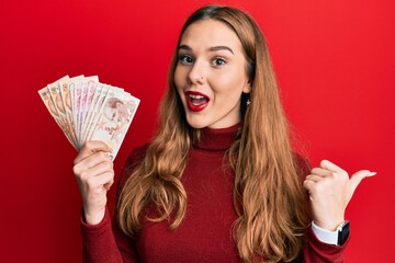 Young blonde woman holding turkish lira banknotes pointing thumb up to the side smiling happy with open mouth