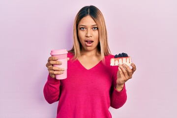 Beautiful hispanic woman holding takeaway cup of coffee and cheesecake in shock face, looking...