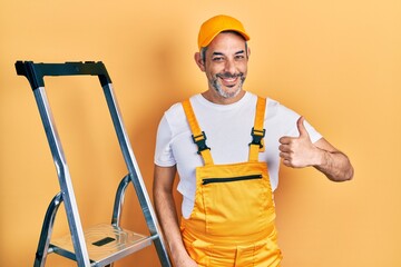 Handsome middle age man with grey hair holding ladder doing happy thumbs up gesture with hand. approving expression looking at the camera showing success.