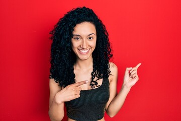 Young hispanic woman with curly hair wearing casual style with sleeveless shirt smiling and looking at the camera pointing with two hands and fingers to the side.