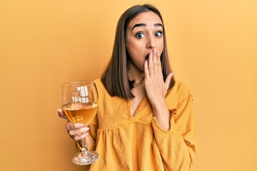 Young brunette girl drinking a glass of white wine covering mouth with hand, shocked and afraid for...