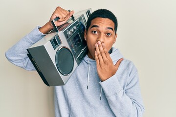 Young handsome hispanic man wearing workout clothes and holding boombox covering mouth with hand,...