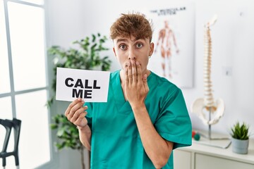 Young caucasian physio man holding call me banner at the clinic covering mouth with hand, shocked...