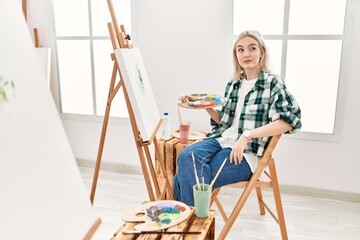 Young artist woman painting on canvas at art studio smiling looking to the side and staring away thinking.