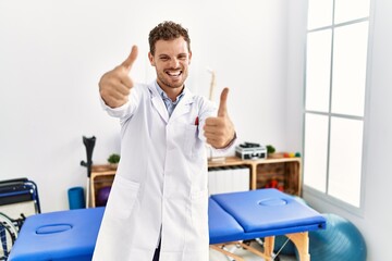 Handsome young man working at pain recovery clinic approving doing positive gesture with hand, thumbs up smiling and happy for success. winner gesture.