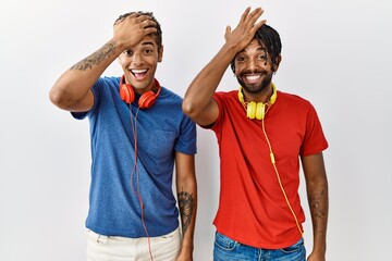 Young hispanic brothers standing over isolated background wearing headphones surprised with hand on...