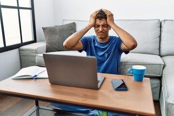 Young handsome hispanic man using laptop sitting on the floor suffering from headache desperate and stressed because pain and migraine. hands on head.