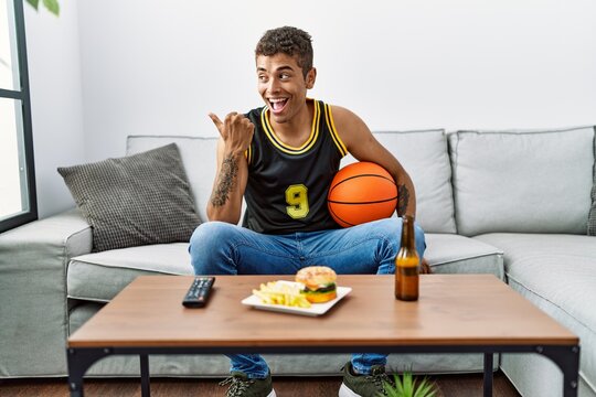 Young handsome hispanic man holding basketball ball cheering tv game pointing thumb up to the side smiling happy with open mouth