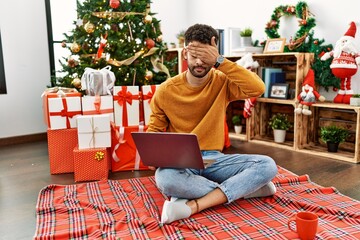 Arab young man using laptop sitting by christmas tree covering eyes with hand, looking serious and sad. sightless, hiding and rejection concept