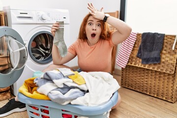 Young redhead woman putting dirty laundry into washing machine surprised with hand on head for mistake, remember error. forgot, bad memory concept.