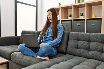 Young chinese girl wearing casual clothes working using laptop at home scared and amazed with open mouth for surprise, disbelief face