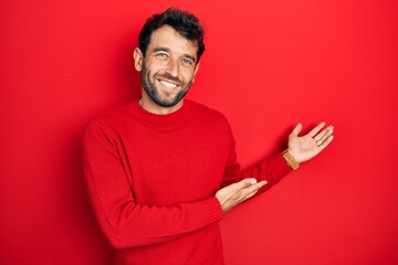 Handsome man with beard wearing casual red sweater inviting to enter smiling natural with open hand