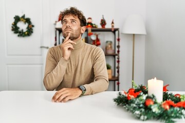 Young handsome man with beard sitting on the table by christmas decoration thinking concentrated...