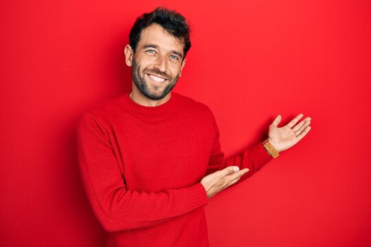 Handsome man with beard wearing casual red sweater inviting to enter smiling natural with open hand