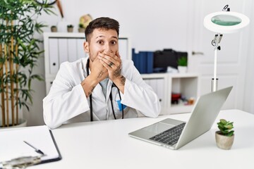 Naklejka premium Young doctor working at the clinic using computer laptop shocked covering mouth with hands for mistake. secret concept.