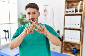 Young physiotherapist man working at pain recovery clinic rejection expression crossing fingers...