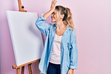 Beautiful young blonde woman standing by white painter easel stand surprised with hand on head for...
