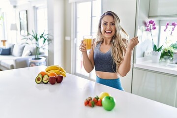 Young caucasian fitness woman wearing sportswear drinking healthy orange juice screaming proud,...