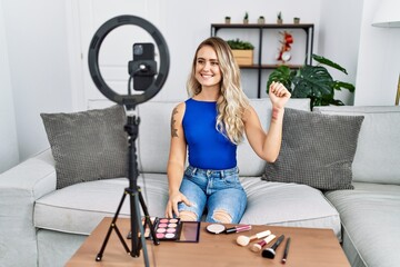Young beautiful woman recording make up vlog tutorial with smartphone at home looking positive and happy standing and smiling with a confident smile showing teeth