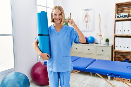 Young caucasian physiotherapist woman holding yoga mat smiling with an idea or question pointing finger with happy face, number one