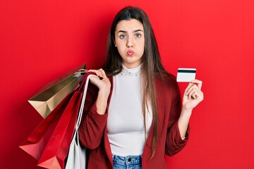 Young brunette teenager holding shopping bags and credit card puffing cheeks with funny face. mouth...