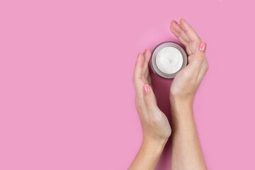 A jar of white cream in the hands of a woman on a pink background. Place for a logo. Face cream or skin care products.