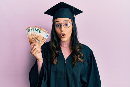 Young hispanic woman wearing graduation uniform holding euro banknotes scared and amazed with open mouth for surprise, disbelief face