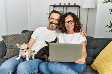 Middle age hispanic couple smiling happy and using laptop. Sitting on the sofa with dogs at home.