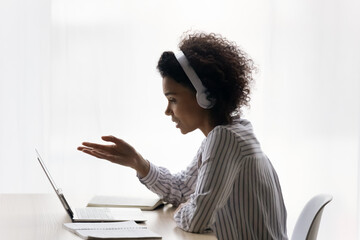 Positive afro american teen female wear earphones sit at desk by laptop learn on distance talk...