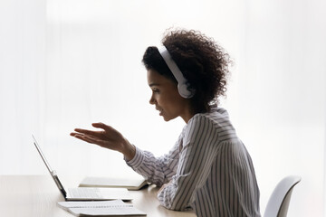 Positive afro american teen female wear earphones sit at desk by laptop learn on distance talk speak ask answer questions at virtual consultation. Black gen z lady confer online in app for video calls