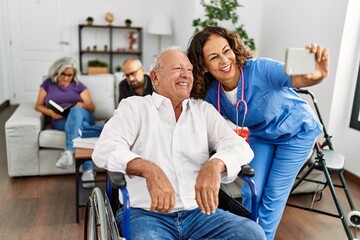 Obraz premium Middle age doctor woman and retired man sitting on wheelchair make selfie by the smartphone at nursing home.