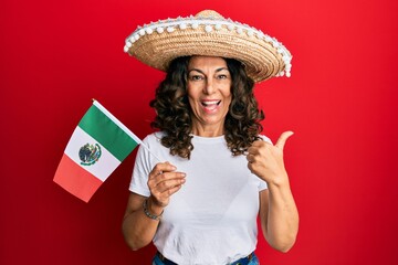 Middle age hispanic woman wearing mexican hat holding mexico flag pointing thumb up to the side smiling happy with open mouth