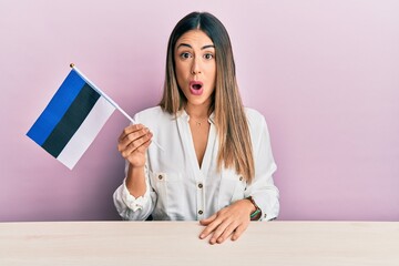 Young hispanic woman holding estonia flag sitting on the table scared and amazed with open mouth for surprise, disbelief face