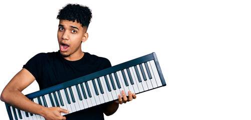 Young african american man holding piano keyboard in shock face, looking skeptical and sarcastic, surprised with open mouth