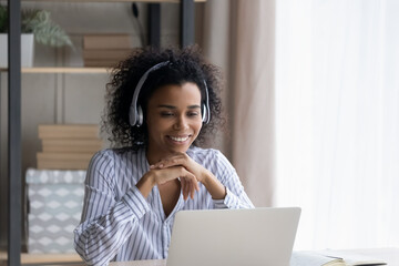 Virtual english lesson. Biracial female sit by desk in headphones watch video class on laptop...