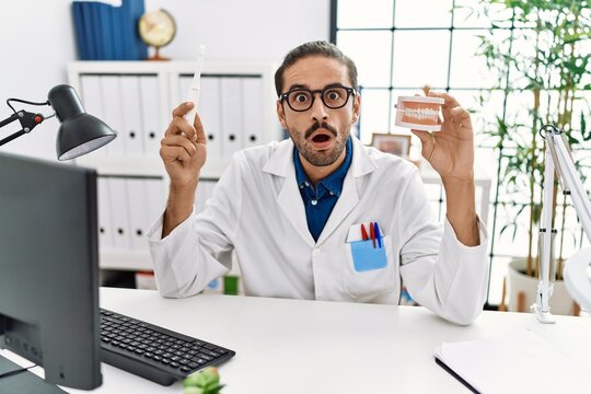 Young hispanic dentist man holding denture and toothbrush afraid and shocked with surprise and amazed expression, fear and excited face.