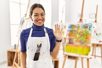 Young brunette woman at art studio waiving saying hello happy and smiling, friendly welcome gesture