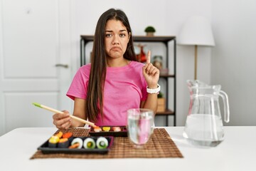 Obraz na płótnie Canvas Young brunette woman eating sushi using chopsticks pointing up looking sad and upset, indicating direction with fingers, unhappy and depressed.