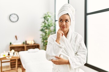 Young brunette woman wearing towel and bathrobe standing at beauty center with hand on chin thinking about question, pensive expression. smiling with thoughtful face. doubt concept.