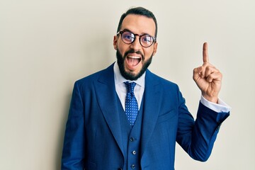 Young man with beard wearing business suit and tie pointing finger up with successful idea. exited and happy. number one.