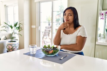 Obraz na płótnie Canvas Young hispanic woman eating healthy salad at home looking confident at the camera with smile with crossed arms and hand raised on chin. thinking positive.