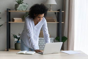 Important news. Thoughtful mixed race female worker stand by office desk read business email on computer pc ponder on electronic document. Young black woman expert look on laptop screen solve problem