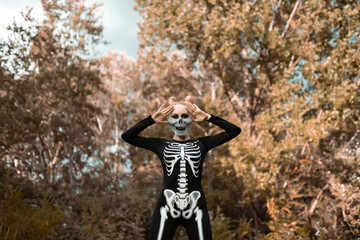 A girl with a scary make-up in a skeleton costume, a mask with rhinestones in a gloomy autumn forest. Stands terribly, looking at the camera. Portrait. Halloween, autumn holiday concept 