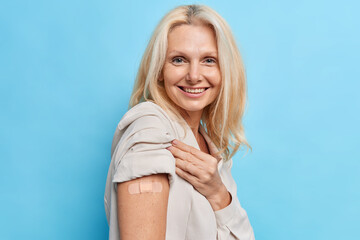 Glad woman shows shoulder with band aid after injection cares about health happy after getting vaccine shot poses against blue studio wall. Disease preventionn coronavirus immunization concept