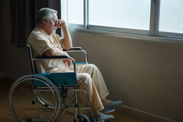 asian senior man patient sit on wheelchair holding head with hands alone and looking out the window...