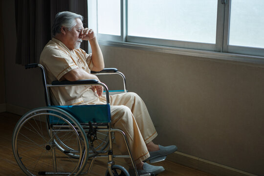 asian senior man patient sit on wheelchair holding head with hands alone and looking out the window at hospital ward . Lonely old male  in Nursing home. loneliness elder. Depressed mature .